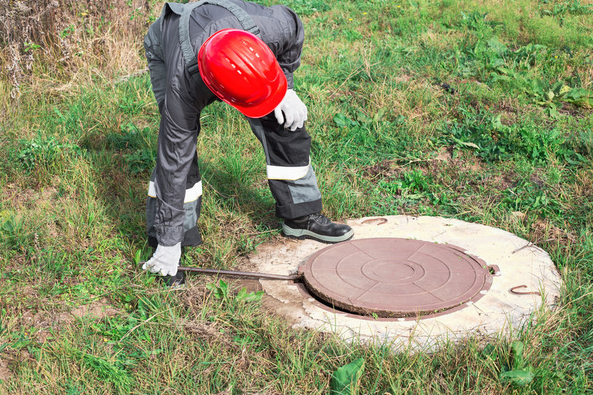A worker in overalls and a helmet opens a manhole with a crowbar. Control and maintenance of septic tank